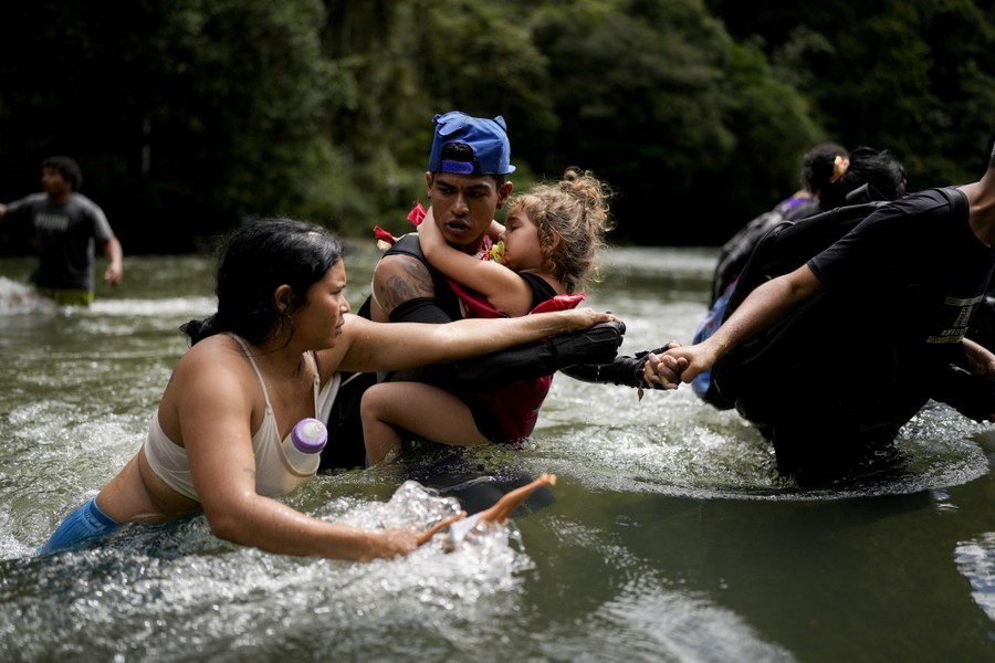 Several people hold hands and help one another cross a waist-deep river.