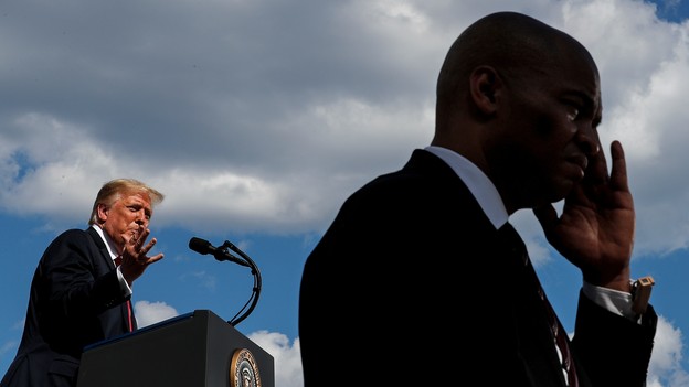 A Secret Service agent in the foreground, with Donald Trump speaking behind him