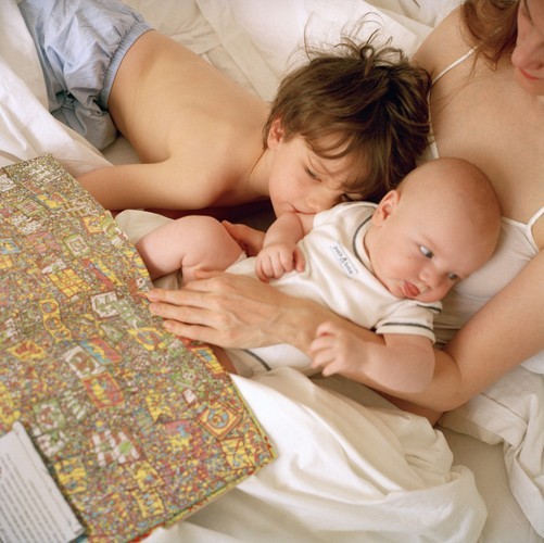A toddler and baby lay in their mother's lap while she reads a book