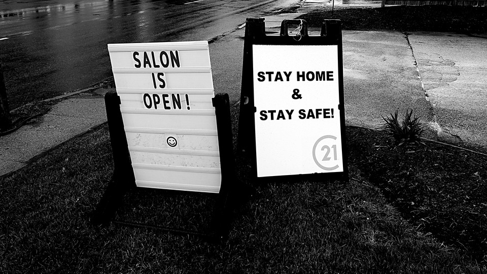 A photo of two road-side signs. One says, "Salon is open!" The other says, "Stay home and stay safe!"