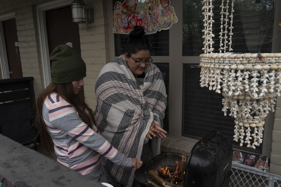 Two women warm themselves beside a small fire in a barbecue on the porch of a house.