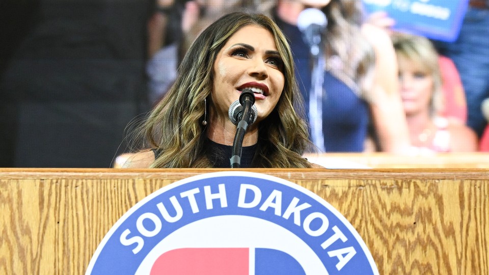 Kristi Noem stands at a podium at a 2023 rally in South Dakota and speaks into a microphone with a crowd behind her