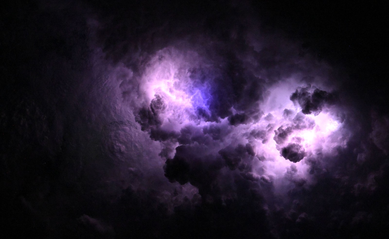 Blue and purple flashes are seen as lightning strikes among cloud formations, seen from above at night.