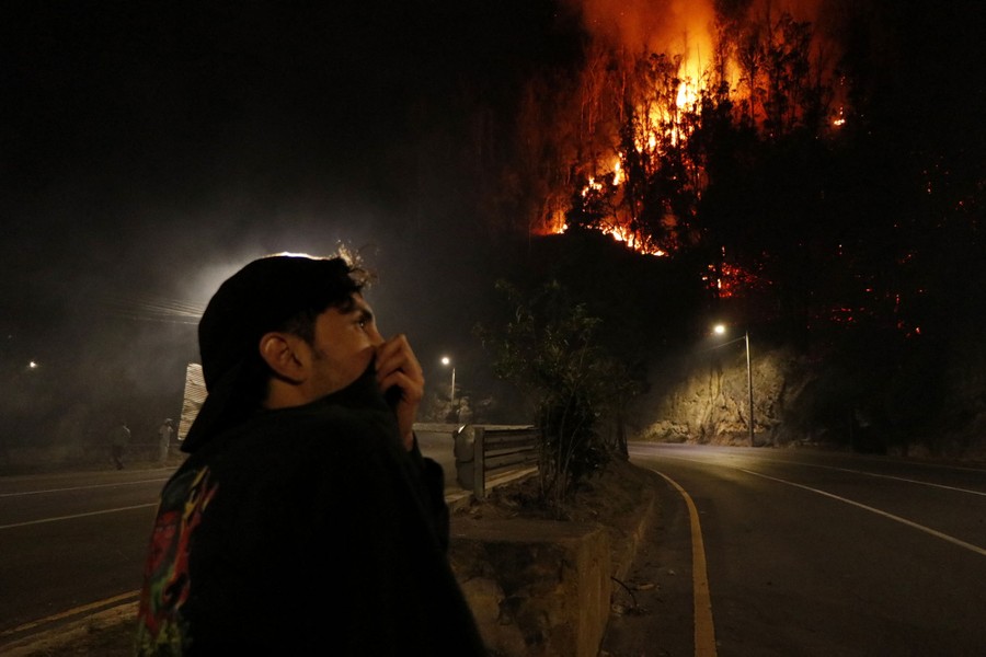 A person stands by the side of a road, covering their face with their shirt, looking up toward a hillside that is on fire.
