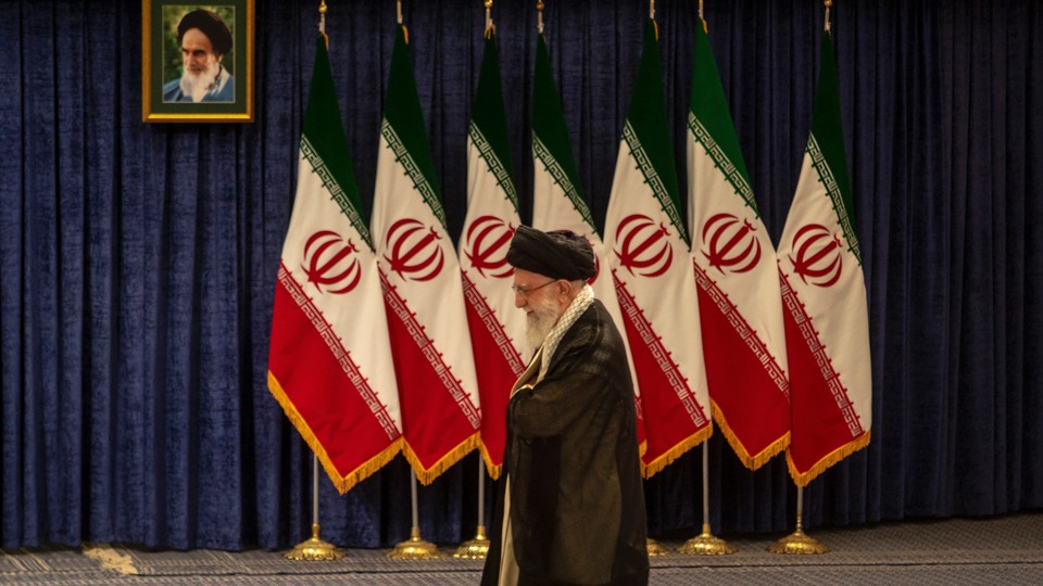 Khamenei, looking down, walks past a bank of seven Iranian flags with a portrait of Ayatollah Ruhollah Khomeini above them.