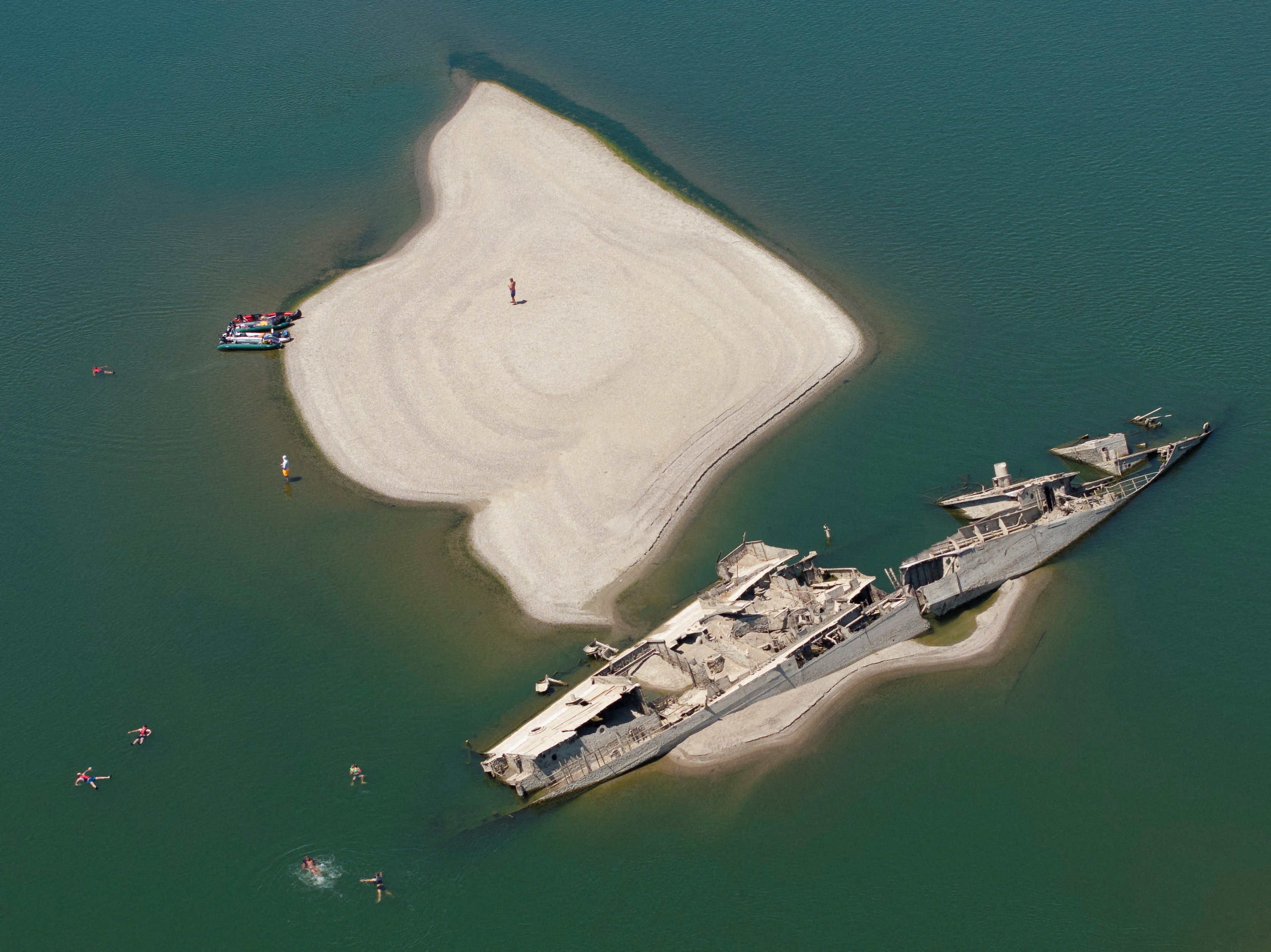A drone view shows people swimming near a sunken WW2-era German warship in a low river.