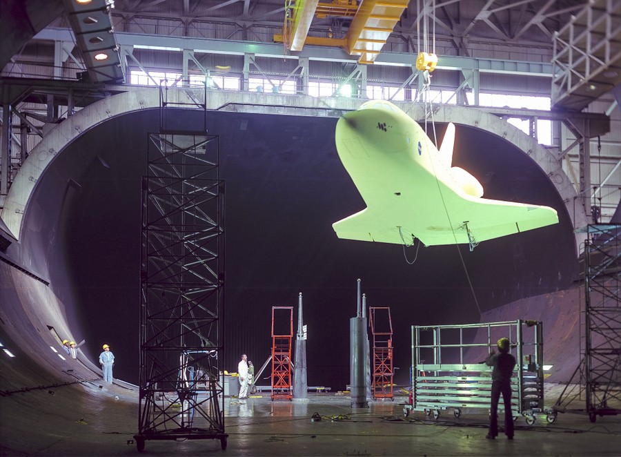 People work inside a large wind tunnel, using a crane to move a huge scale model of the space shuttle.