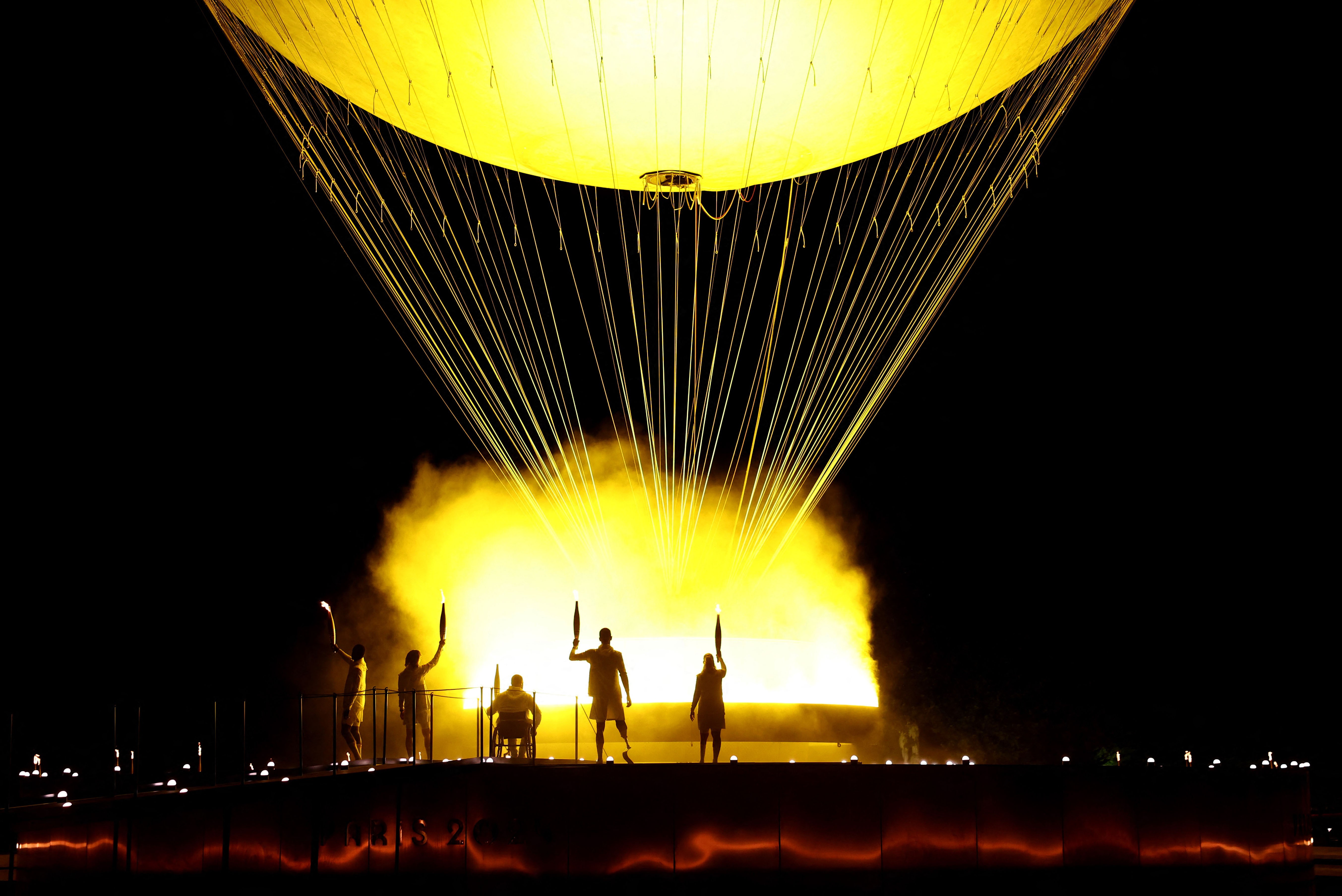 Five torchbearers stand beside a large, gondola-like cauldron, beneath a large balloon, its silhouette standing out against the glowing smoke.