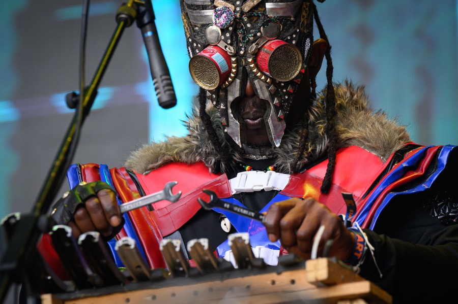 A masked performer plays a handmade percussion instrument using wrenches as mallets.