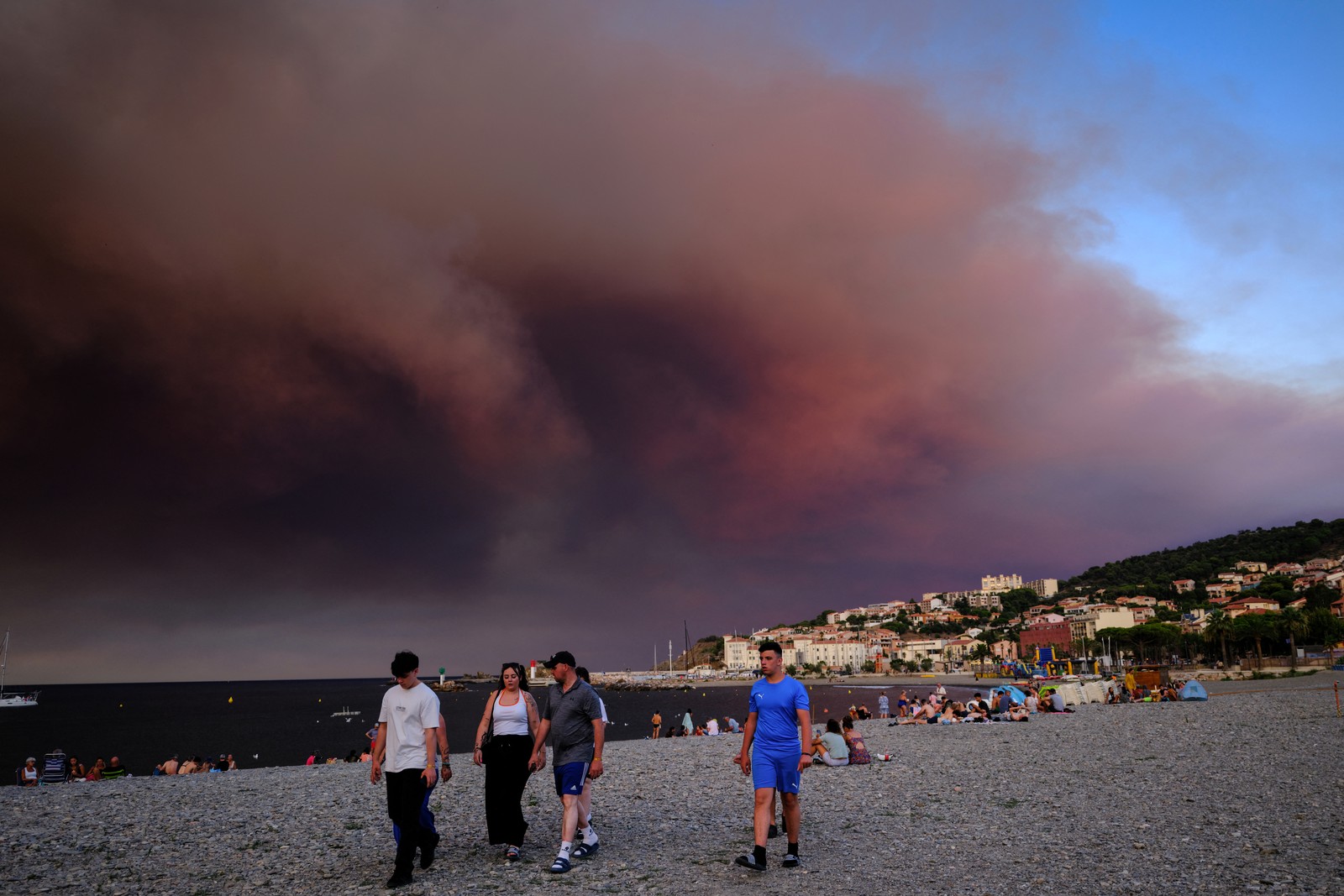 Tourists walk on a beach as dark smoke from wildfires rises above them.