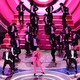 Ryan Gosling, dressed as Ken in a pink suit and gloves, sings into a microphone on the Oscars stage as several dozen men in black tuxedos dance behind him.