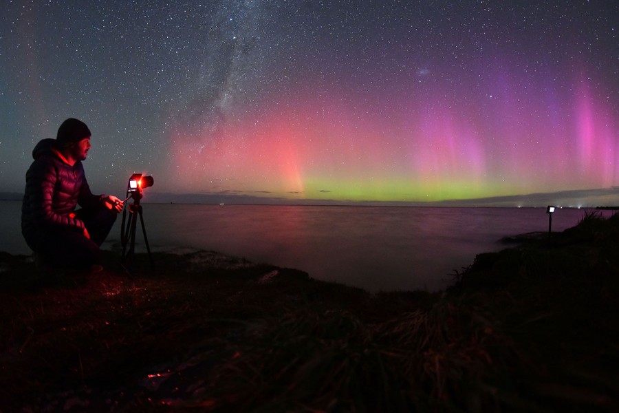 A person points a camera on a tripod toward the horizon at night, beneath the glowing red and purple lights of an aurora.