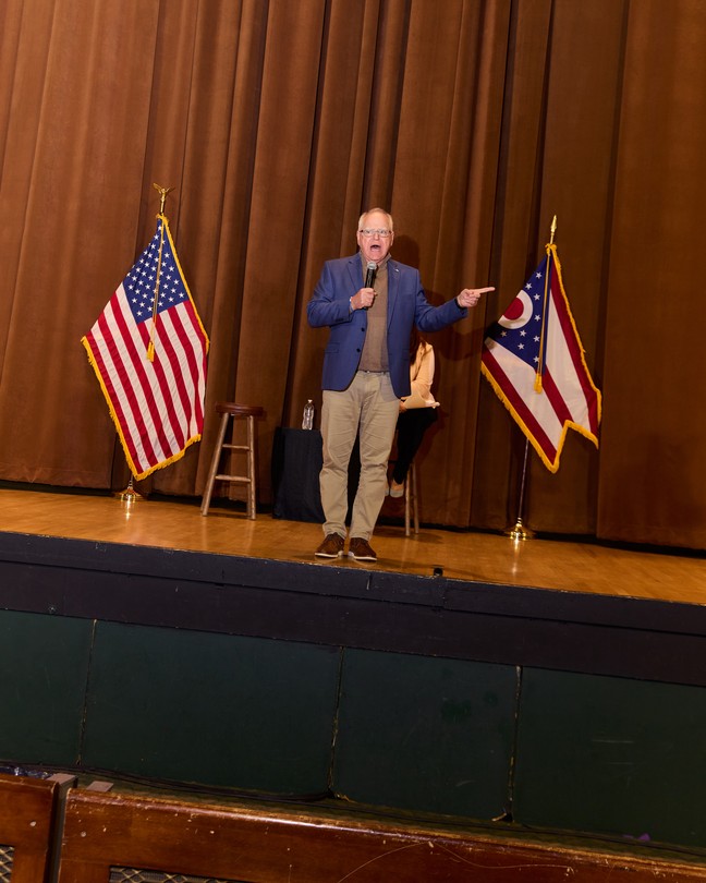 Tim Walz standing on a stage between an American flag and an Ohio flag