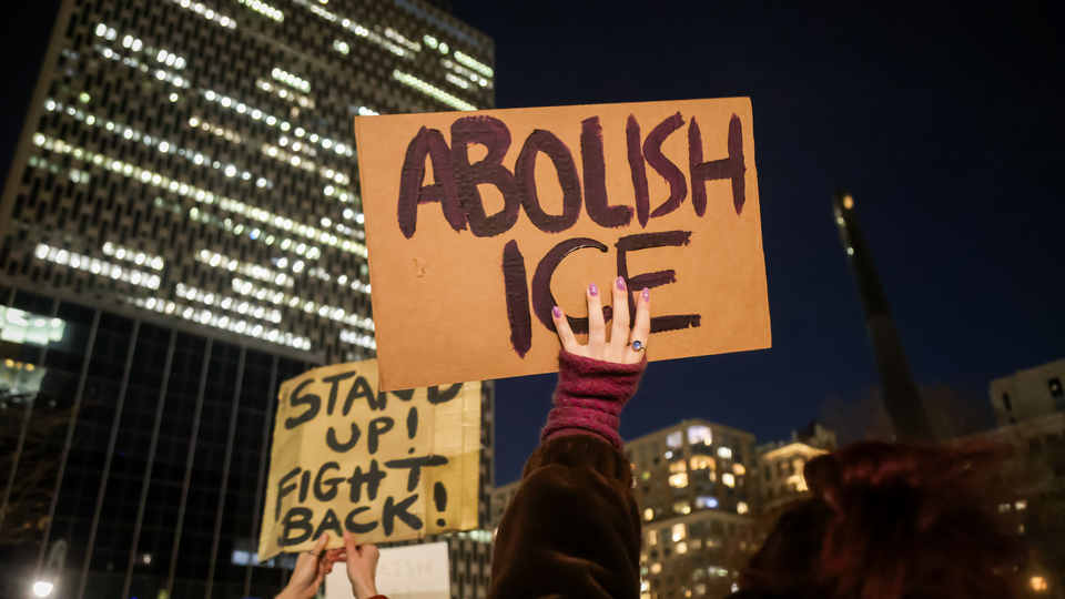 Protestors hold up signs reading "ABOLISH ICE" and "STAND UP! FIGHT BACK!" against a backdrop of a dark city and lit-up buildings.