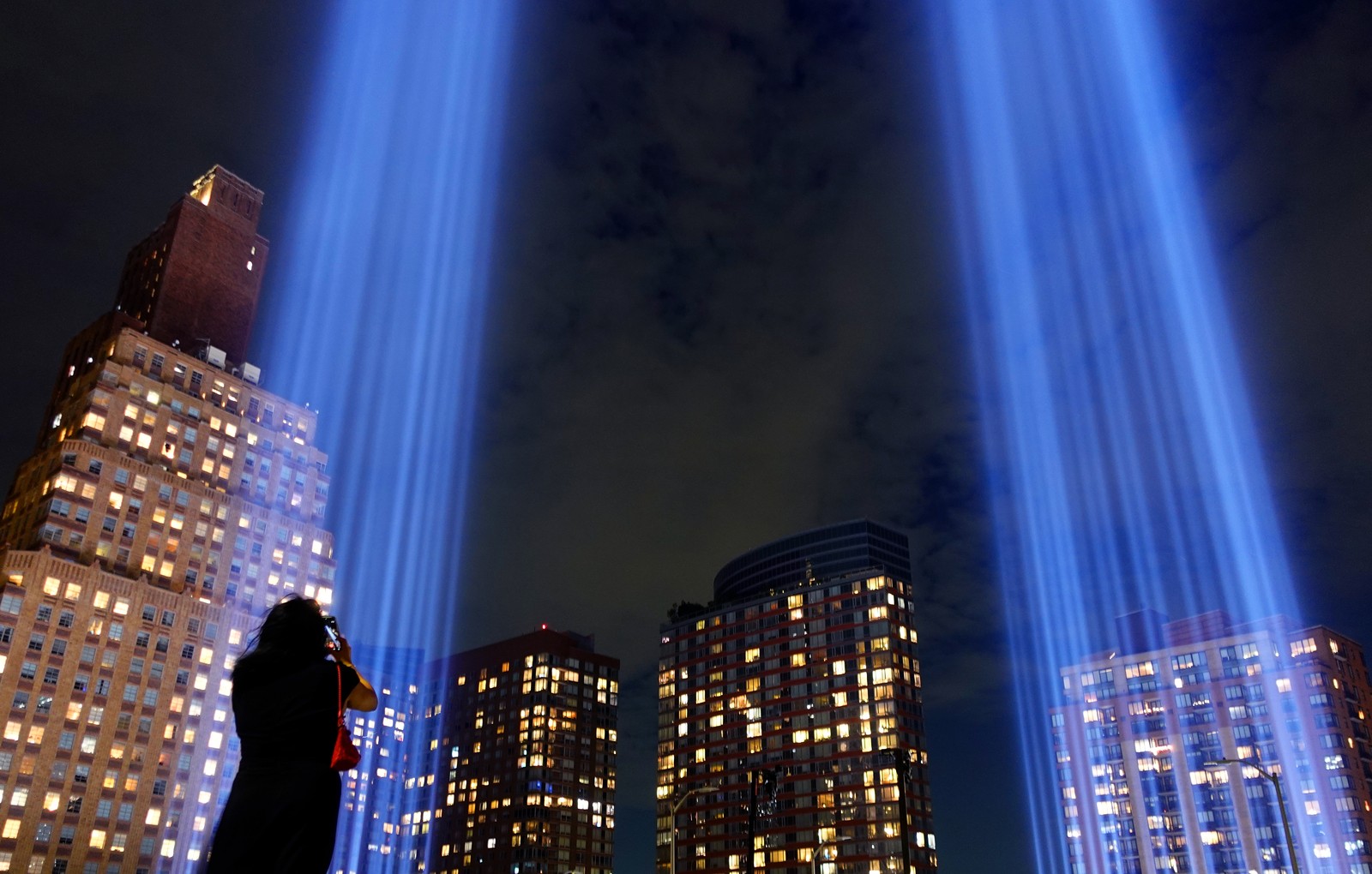 A woman looks upward, taking a photograph of two broad and bright beams of light reaching into the sky in New York City.