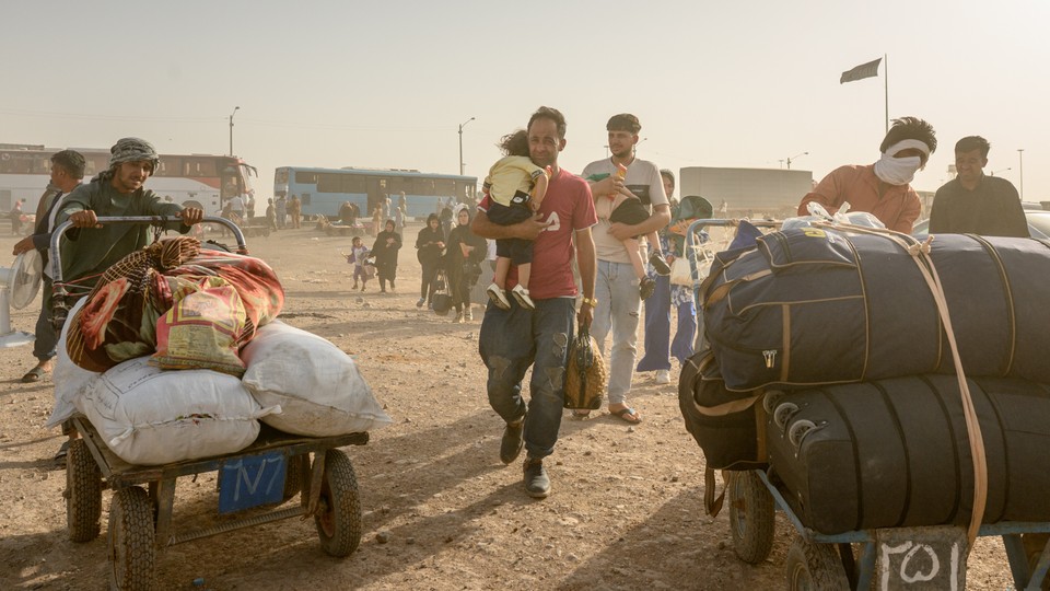 Photograph showing crowd of migrants walking with carts of their belongings; man holding small child