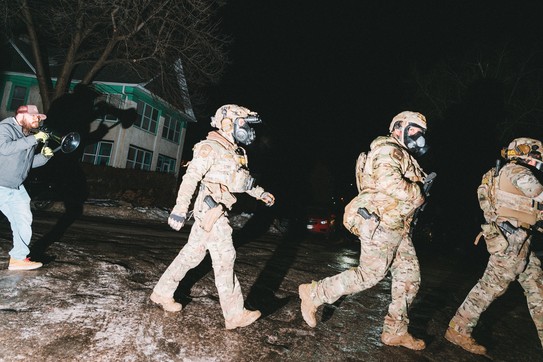 A man with a megaphone chases three ICE agents in a neighborhood at dusk