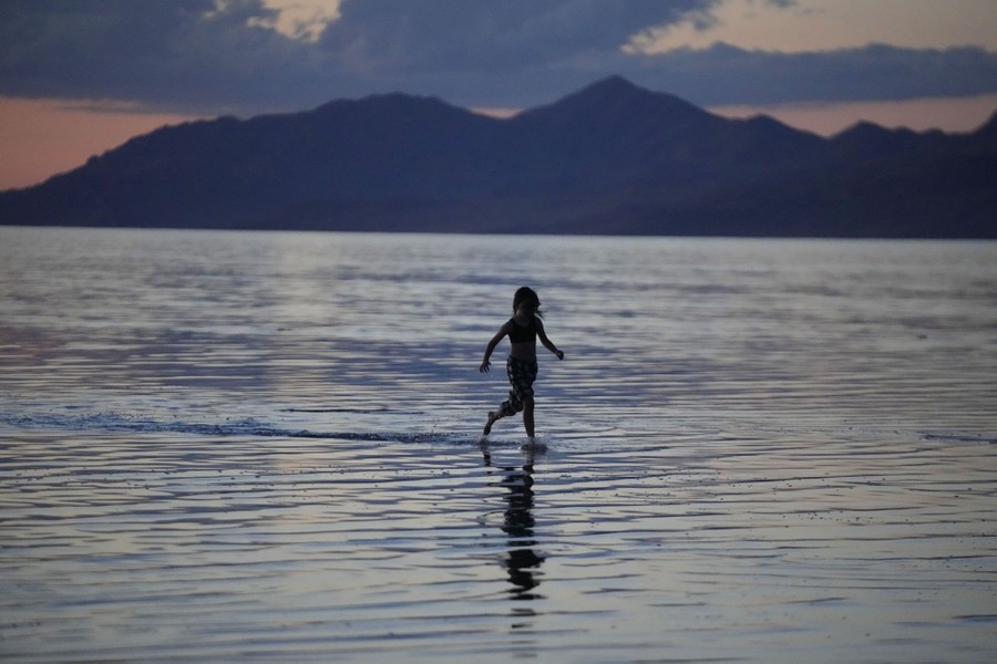 A young person runs through shallow water, with a broad lake and mountains visible in the background.