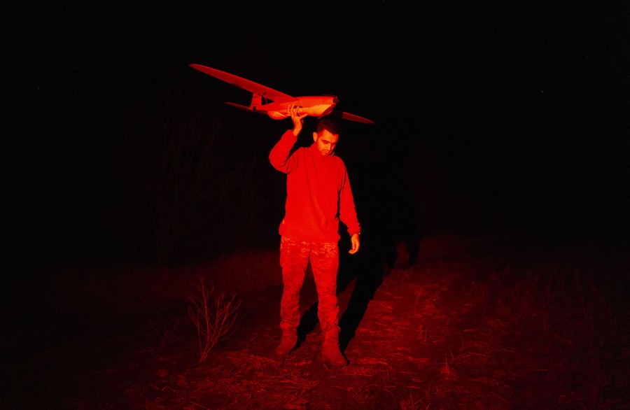 A soldier lit by red light holds an airplane-shaped drone above his head at night, preparing to launch it.