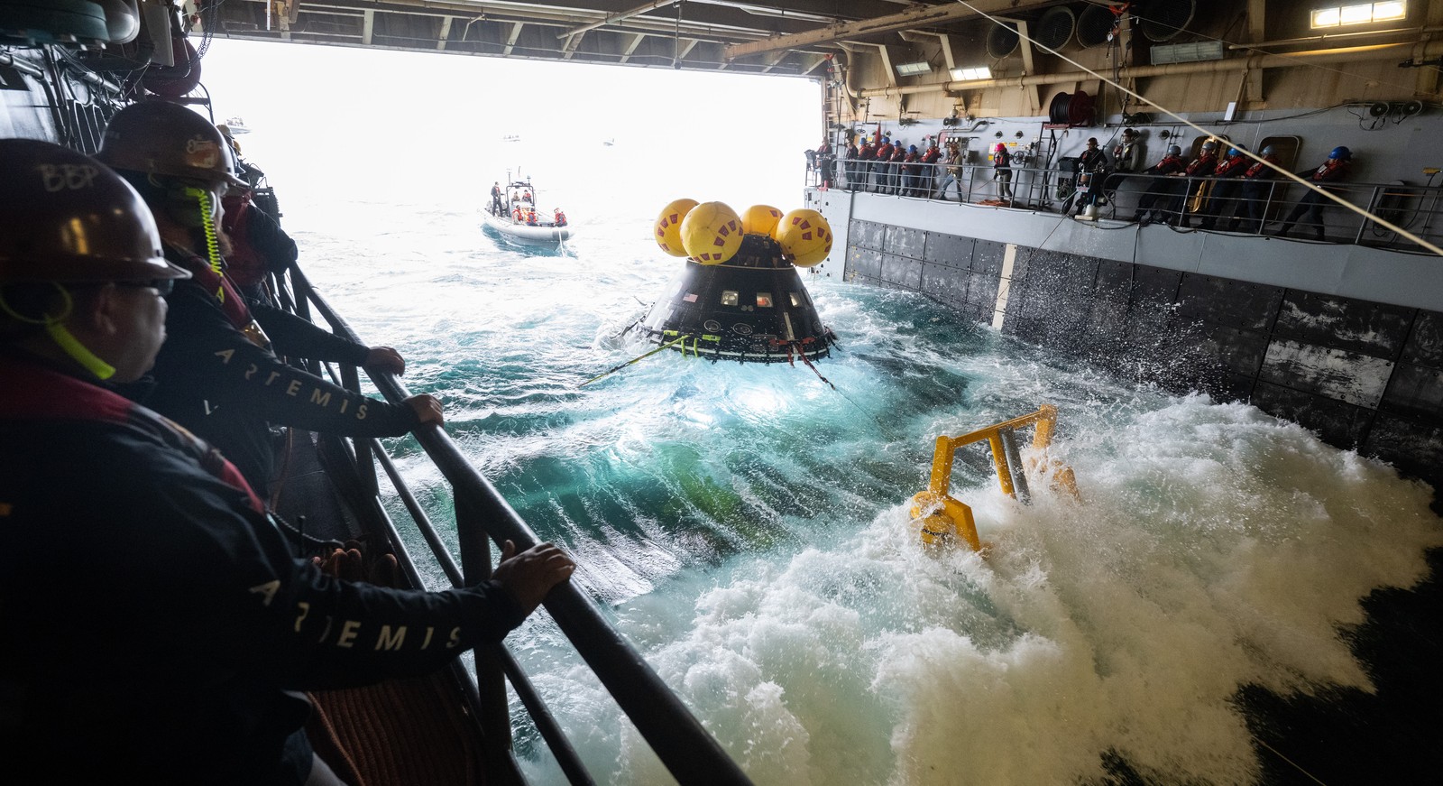 People stand on decks inside an open space at the bottom of a ship, on either side of a space capsule bobbing in waves.