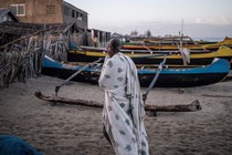 A man on the beach stands near several different-colored canoes.