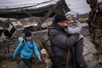 A photo of a man with a gun slung behind his back carrying a child through rubble and a girl in a blue jacket walking behind them. A dog follows her. A soldier in military fatigues stands nearby.