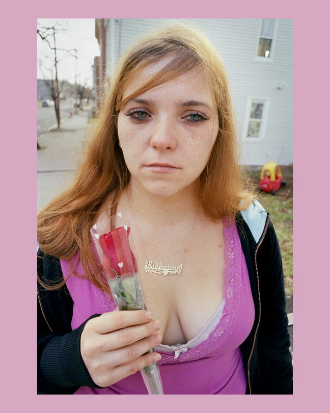 A woman holds a rose in plastic on the street looking at the camera.