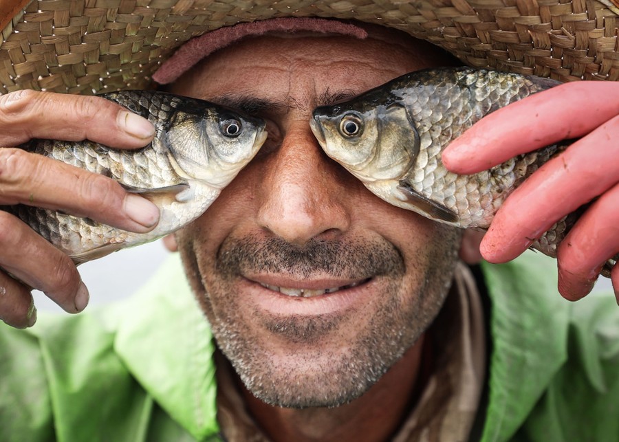 A fisherman holds up two fish, making a sort of a mask, with the eyes of the fish in place of his own.