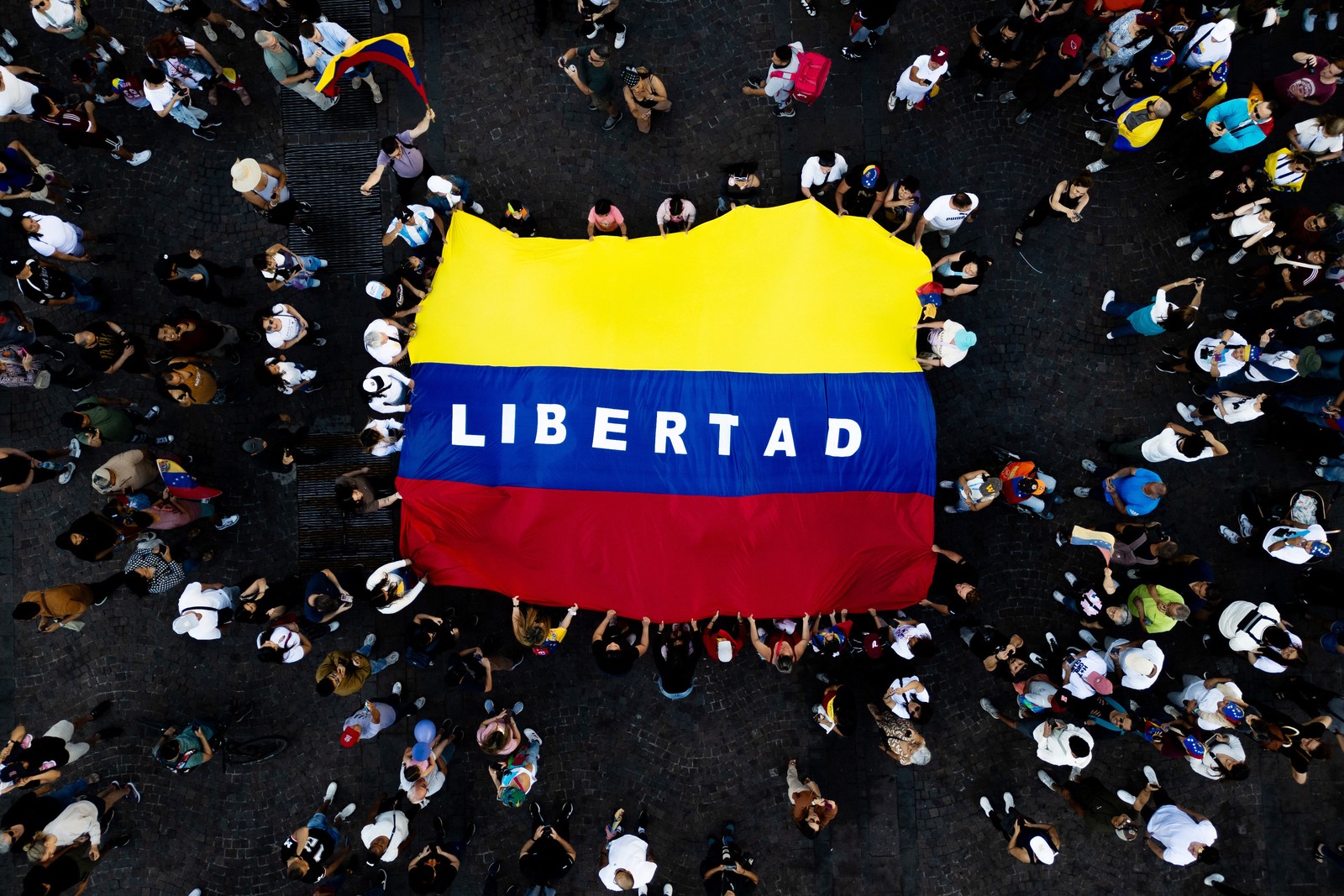 A top-down view of people gathered in a public square who are holding up a large yellow blue and red flag with the word 