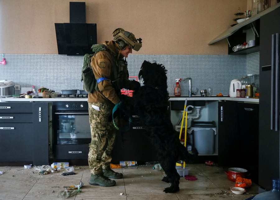 A soldier stands in an abandoned kitchen playing with a dog who is standing on its hind legs in front of him.