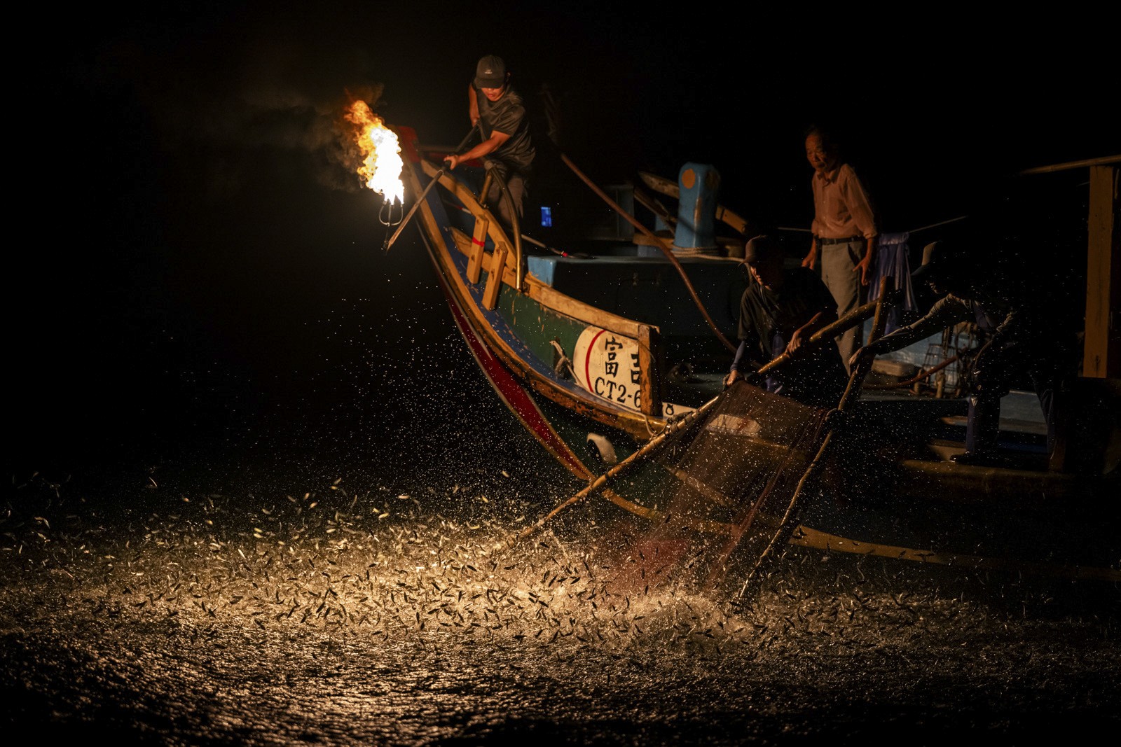 Several people stand on a the deck of a fishing boat at night. One holds a torch aloft, while hundreds of small fish leap from the water's surface below.