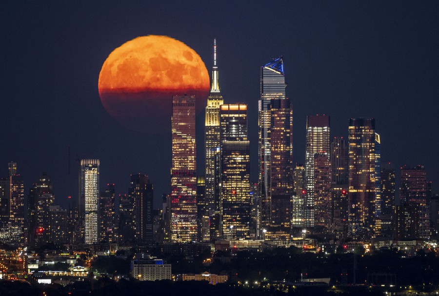 A full moon, partially blocked by clouds, rises behind the New York City skyline.
