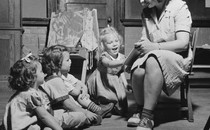 Black-and-white photo of a woman sitting in a chair reading a storybook to three kids seated on the floor