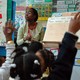 A teacher sits in front of her class. Students raise their hands.