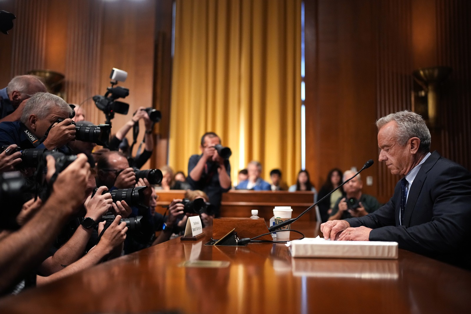 A group of photographers leans in to take pictures of Robert F. Kennedy Junior, who sits at a table before a Senate committee.