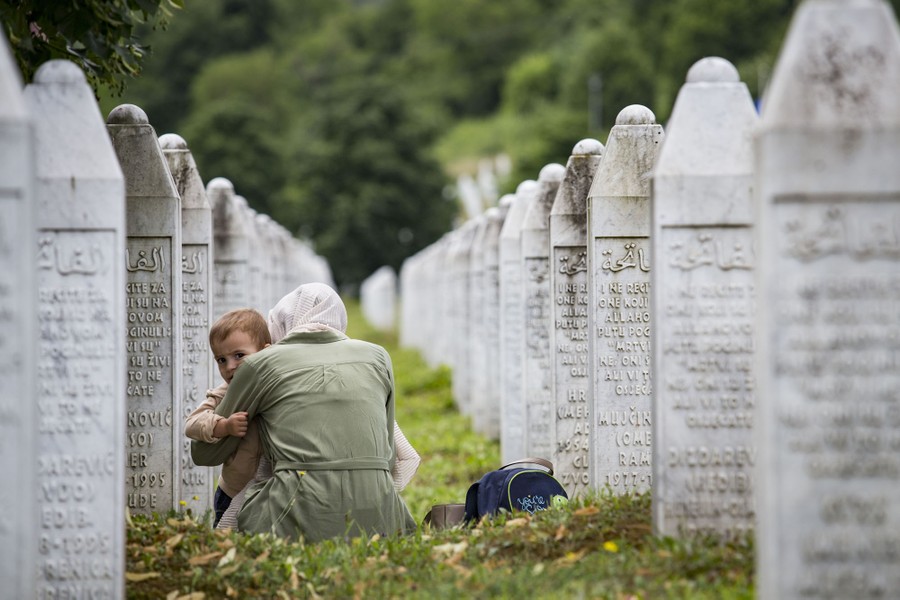 A woman holds a child while sitting on the ground among gravestones.