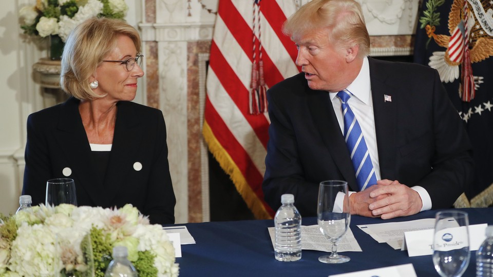 President Donald Trump sits at a table next to Education Secretary Betsy DeVos. 