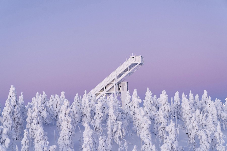 The top of a ski jump stands above a group of snow-covered trees.