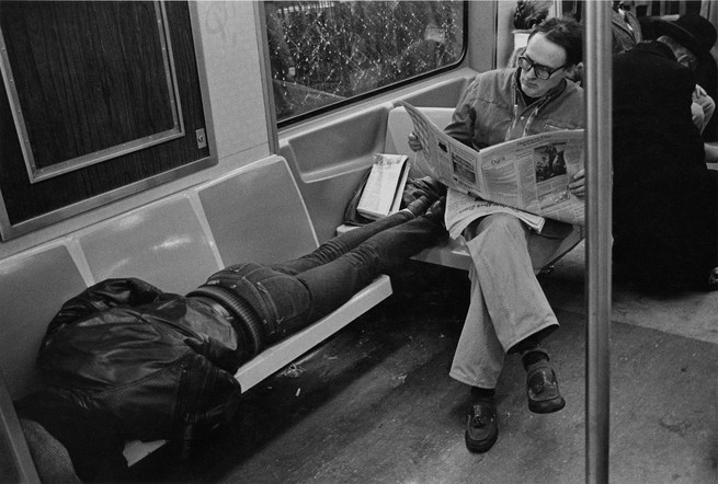 a man reads a newspaper on the subway sitting with another man's feet who is sleeping across benches.