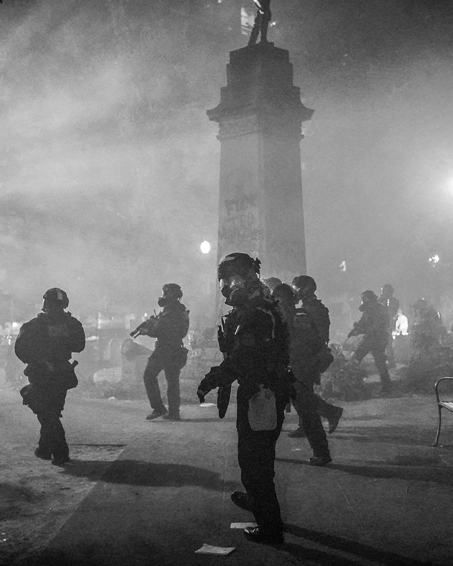 Fotografía en blanco y negro de soldados armados con protección completa y máscaras antigás en un parque brumoso de la ciudad, cerca de una estatua en un pedestal alto.
