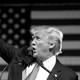 A black-and-white photo of President Donald Trump shows him waving as he stands behind a podium.