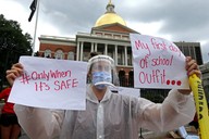 A woman wearing a face shield, surgical mask, and plastic poncho holds up signs in protest of school reopening.