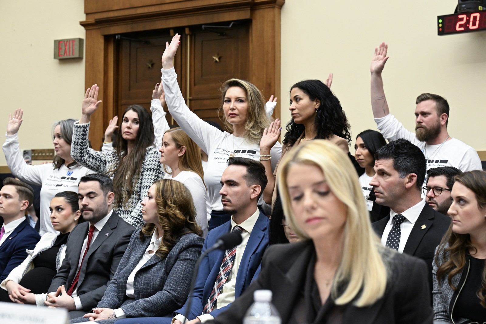 People in the back of a congressional hearing room stand and raise their hands.