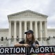 A person in a black cap stands in front of a columned building, behind two signs that each read "Abortion."
