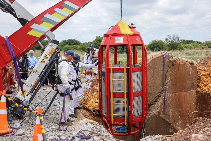 Workers stand beside a rescue cage suspended above a mine shaft.