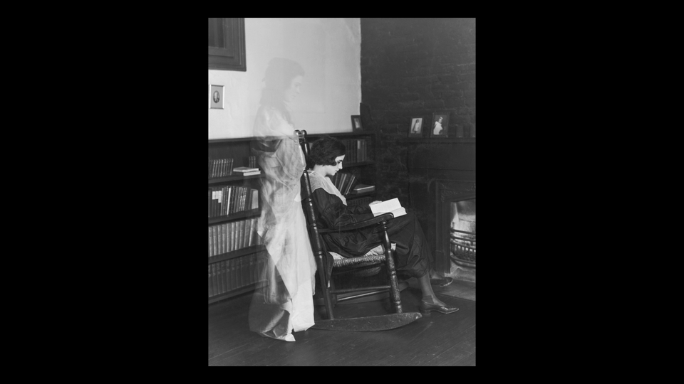 A black and white photograph of a woman reading in a rocking chair, with a ghostly figure of another woman standing behind her looking over her shoulder