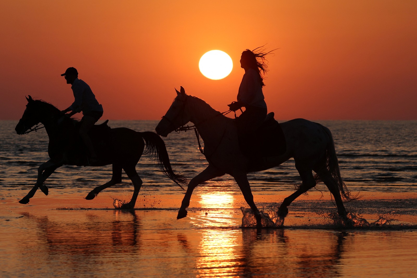 People ride horses on a beach at sunset.