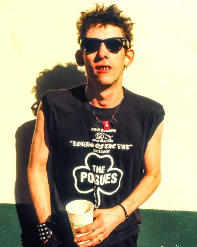 Shane MacGowan standing against a cream and green wall wearing a Pogues black shirt and sunglasses