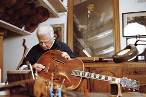 photo of man with white hair sitting behind desk and holding acoustic guitar and surrounded by other musical instruments, with large antique portrait of man playing violin in background