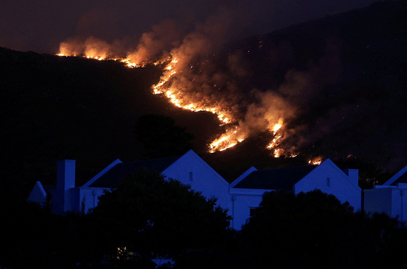A line of a wildfire burns along a hillside, seen behind houses at night.